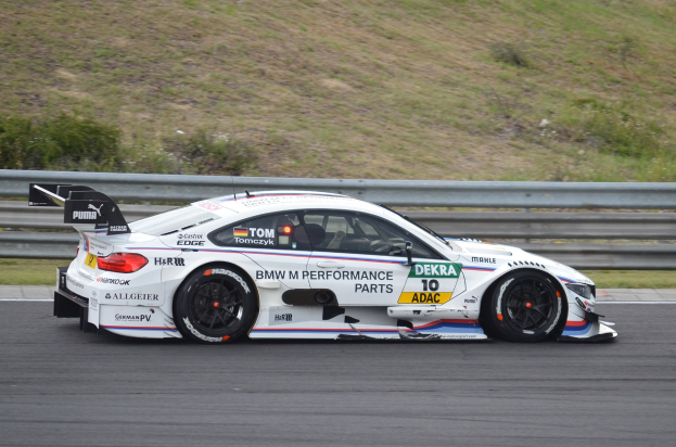 BMW M4 DTM-Rennwagen auf einer Rennstrecke mit Sponsorentext, umgeben von grünem Gras und einer Absperrung im Hintergrund.