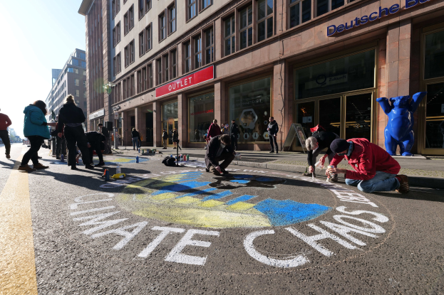 Menschen sitzen vor einem Gebäude mit Fenstern und Schildern auf dem Boden, umgeben von Flaschen und anderen Gegenständen, während sie an einer Klimaprotestaktion in Berlin teilnehmen, bei der es einen klaren blauen Himmel gibt.