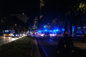 Eine nächtliche Straßenansicht einer Stadt mit Polizeiwagen und Bussen, die fahren, Menschen auf dem Bürgersteig gehen, Gras auf der linken Seite, Bäume und Gebäude im Hintergrund und Laternen, die die Straße beleuchten.