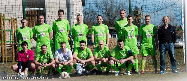 Eine Gruppe von Männern in grünen T-Shirts steht auf einem Fussballfeld, einige hocken, mit einem Ball vor ihnen in der Nähe eines Torpfostens; Bäume, Gebäude und Fahrzeuge sind im Hintergrund zu sehen.