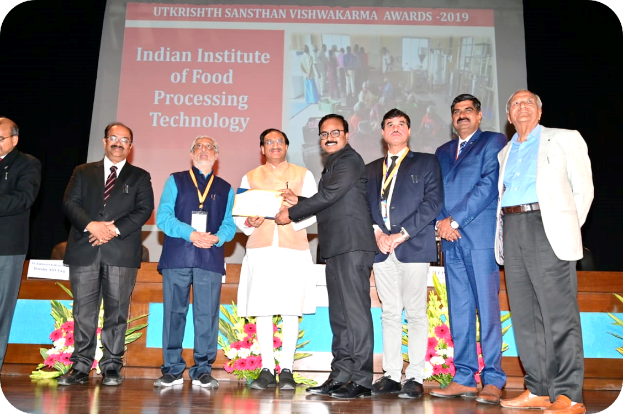 Eine Gruppe von Männern steht auf einer Bühne, zwei halten ein Zertifikat, mit einem Bildschirm im Hintergrund, der 'Indian Institute of Food Processing Technology Awards 2019' anzeigt, und Blumenschmuck auf der Bühne.