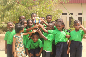 Gruppe von Kindern in einer Schulumgebung mit einem Pokal, Bäumen, Pflanzen und einem Gebäude im Hintergrund.