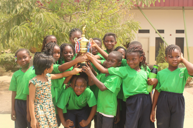 Gruppe von Kindern in einer Schulumgebung mit einem Pokal, Bäumen, Pflanzen und einem Gebäude im Hintergrund.