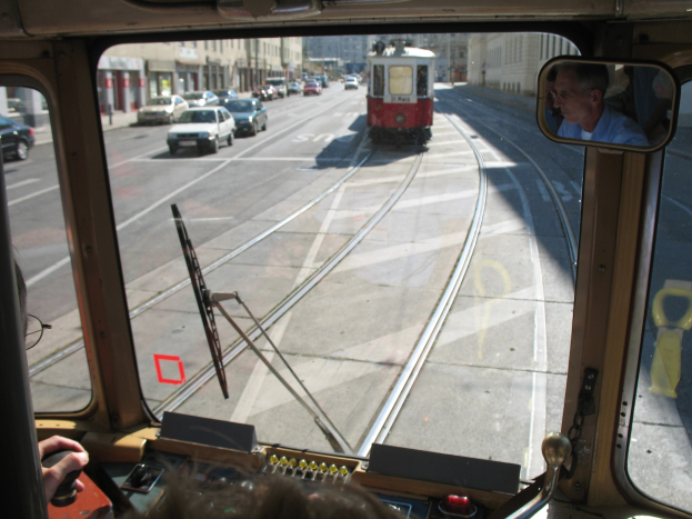 Ein Mann fährt einen Tram auf einer Stadtstraße mit anderen Fahrzeugen, Gebäuden und einem klaren blauen Himmel im Hintergrund.