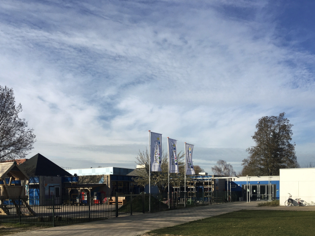 Großes Gebäude mit Metallzaun, Rasenfläche, Weg, Bäumen, Schildern, einem abgestellten Fahrrad, nahen Häusern und einem bewölkten Himmel, identifiziert als der neue Spielbereich für Kinder an der Schule.