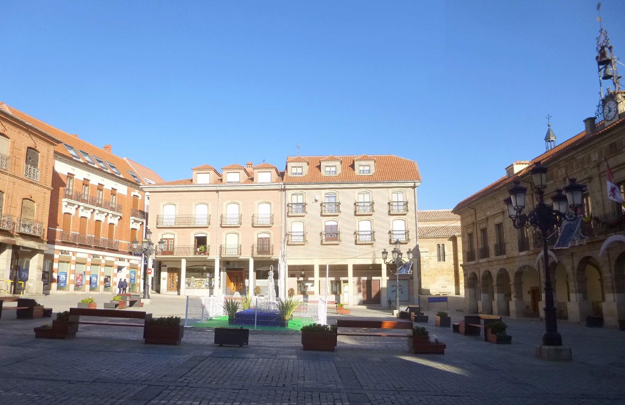 Ein Stadtplatz mit einem zentralen Brunnen, umgeben von Bänken, Topfpflanzen, Straßenlaternen und -pfosten, mit einem Uhrenturm und Gebäuden im Hintergrund unter einem klaren blauen Himmel.
