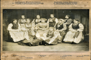 Eine Gruppe von Frauen in traditioneller russischer Kleidung posiert für ein Foto, wobei einige auf dem Boden sitzen und andere stehen, vor einem Vorhanghintergrund, mit Text oben und unten auf dem Bild.