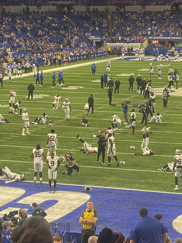 Ein Fußballspiel wird in einem Stadion mit stehenden Zuschauern und einigen auf Stühlen sitzenden Personen sowie einer Person mit einer Kamera für die Live-Übertragung gespielt.