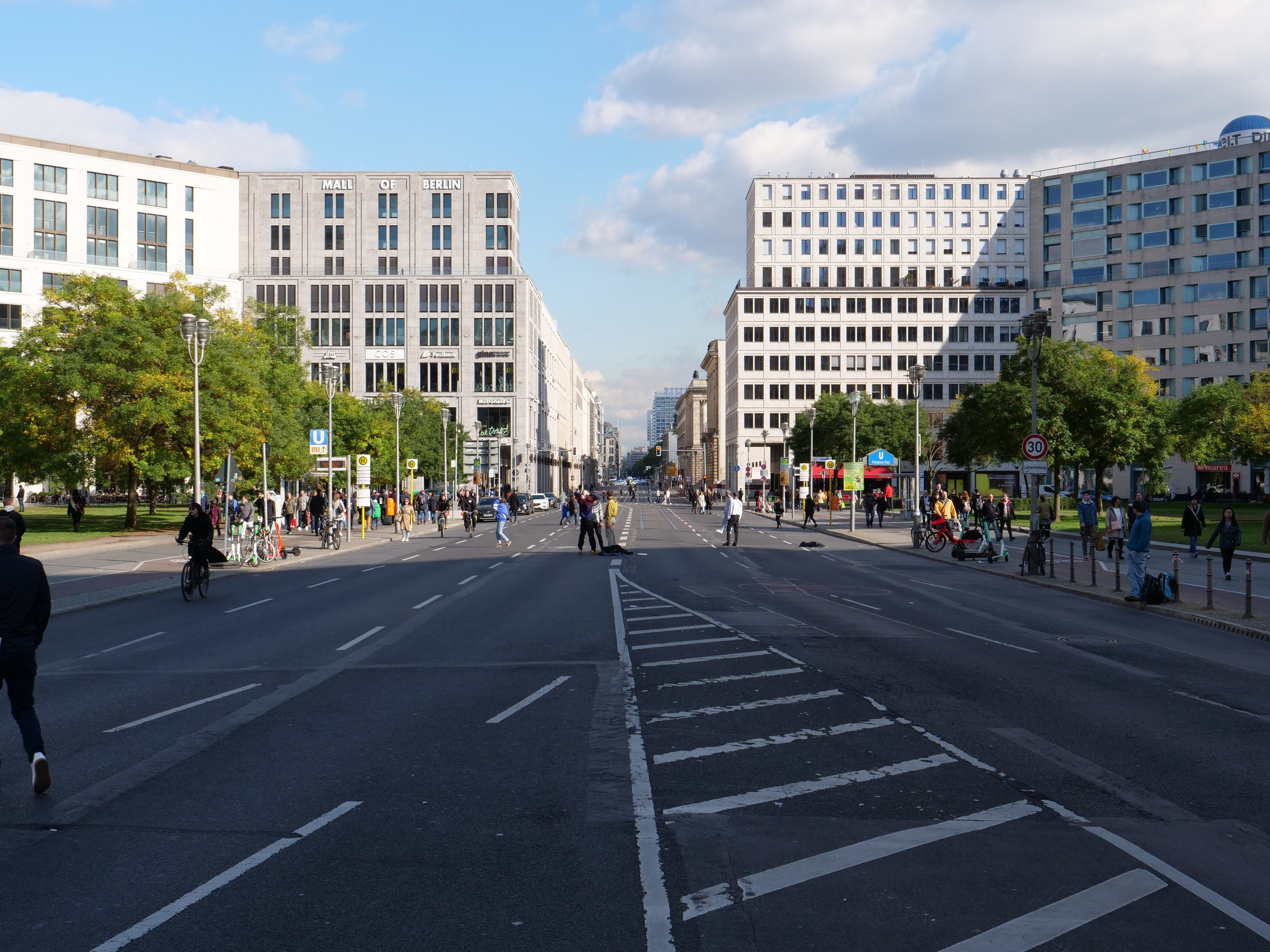 Eine belebte Stadtstraße in Berlin, Deutschland, mit Fußgängern und Radfahrern auf der Straße, hohen Gebäuden mit Fenstern, Bäumen, Laternen und Schildern, unter einem bewölkten Himmel.