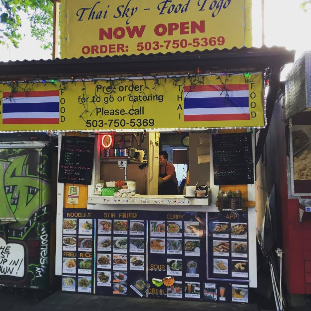 Ein Schaufenster mit verschiedenen Artikeln wie Getränkedosen, Speisekarten, einer Uhr, Gewürzen in Plastikbehältern, Kondimente, Werbetafeln und Namensschildern unter einem Himmel.