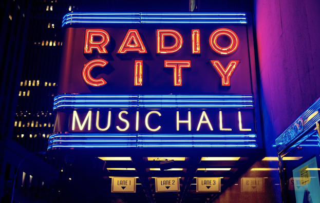Außenansicht des Radio City Music Hall in New York City bei Nacht, hell erleuchtet mit hohen Gebäuden im Hintergrund, mit einer 'Radio City'-Leuchtschrift und zusätzlichen Schildern auf der rechten Seite.