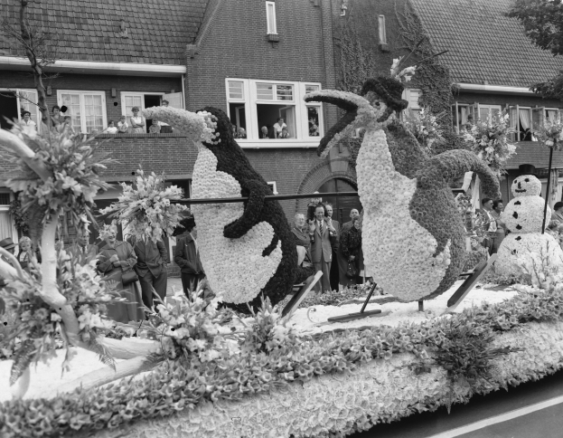 Schwarz-weiß-Foto eines Paradewagens, der mit Blumen und Pinguinfiguren geschmückt ist, mit Menschen auf der Straße und Gebäuden mit Fenstern im Hintergrund.