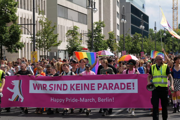 Eine Gruppe von Menschen marschiert bei einer Pride-Veranstaltung in Berlin, die eine pinkfarbene Fahne mit der Aufschrift 'Happy Pride March Berlin' hält, einige tragen Schirme und Fahnen, vor einem Hintergrund aus Laternenmasten, Bäumen, Gebäuden, einem Turm und einem klaren blauen Himmel.
