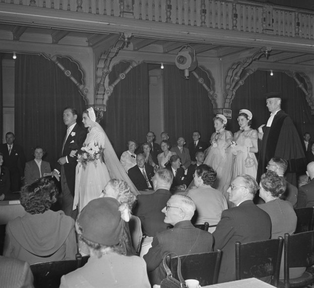 Ein Schwarz-Weiß-Foto einer Hochzeitszeremonie in einem großen Raum mit vielen Gästen, einige halten Blumensträuße, und Tische mit Tassen und anderen Gegenständen, mit Vorhängen und Deckenlampen im Hintergrund.