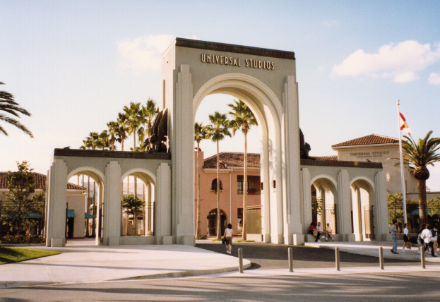 Eingang zu Universal Studios Hollywood mit einem Bogen, einer Statue, Menschen, Grünfläche, Gebäuden, einem Fahnenmast mit einer Fahne und einem bewölkten Himmel.