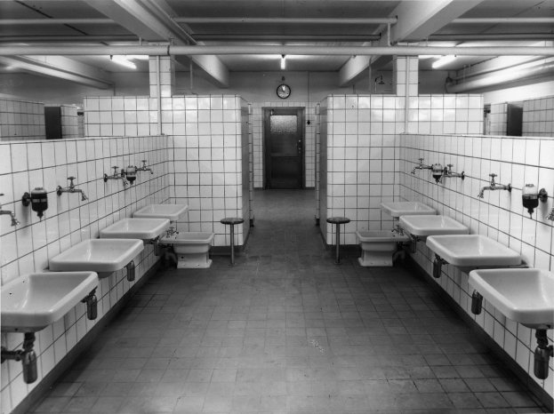 Schwarz-weiß-Foto einer öffentlichen Toilette mit Waschbecken, Hähnen, Hockern, einer Wanduhr und Deckenleuchten.