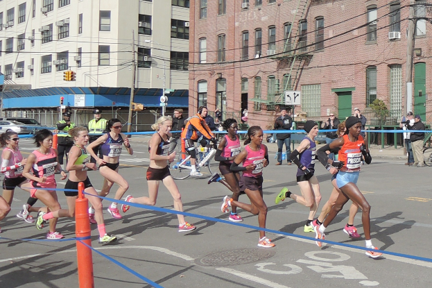 Gruppe von Menschen, die bei einem Marathon auf einer Straße mit Gebäuden, Strommasten, Verkehrszeichen, Fußgängern, einem Radfahrer, Bäumen und einem klaren blauen Himmel laufen.