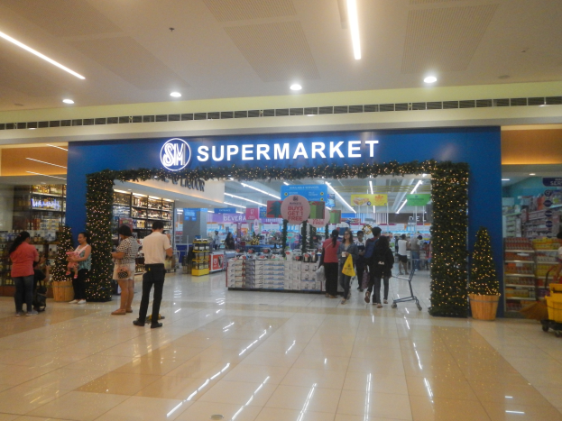 Eine Gruppe von Menschen vor einem Supermarkt, einige halten Einkaufstaschen, mit Einkaufswagen, Topfpflanze, Regalen, Schildern und Deckenbeleuchtung im Bild.