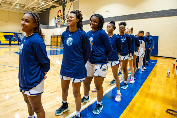 Eine Gruppe junger Frauen in blauen T-Shirts auf einem Basketballfeld, die lächeln, mit Flaschen und Gegenständen auf der rechten Seite und Zuschauern im Hintergrund.