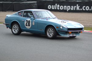 Blauer Wagen auf Rennstrecke mit Helmträger am Steuer, umgeben von Reifen, einer Fahne und Graslandschaft.