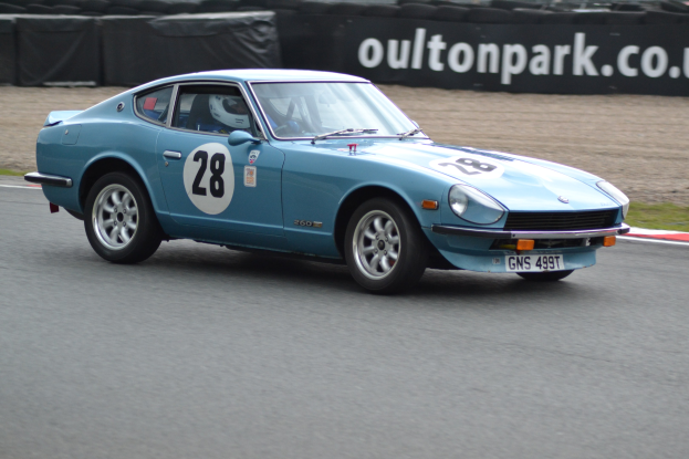 Blauer Wagen auf Rennstrecke mit Helmträger am Steuer, umgeben von Reifen, einer Fahne und Graslandschaft.
