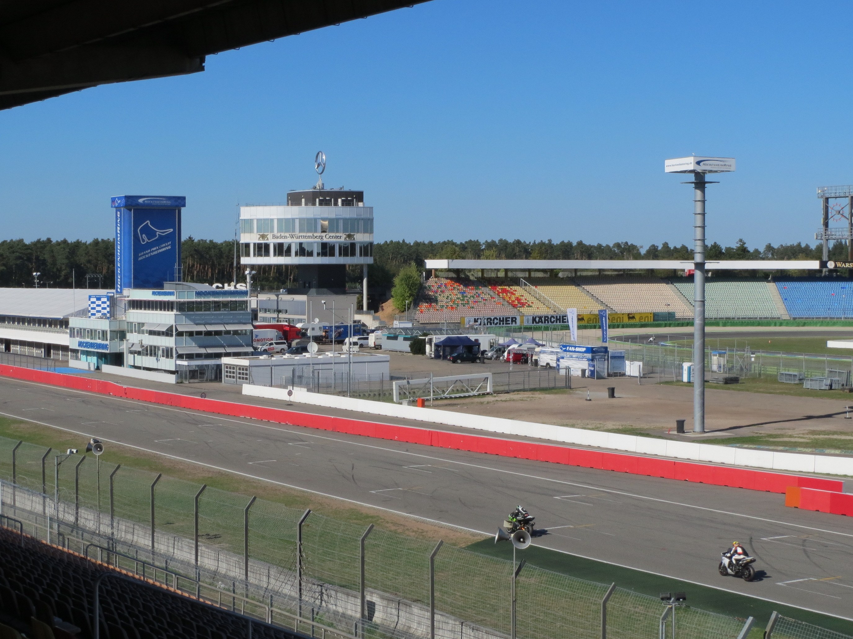Eine Gruppe von Menschen auf Motorrädern auf einer Rennstrecke, umgeben von einem Zaun, Fahrzeugen, einem Gebäude, Schildern, Pfosten, einem Stadion, Beleuchtung, Bäumen und einer bewölkten Himmel.
