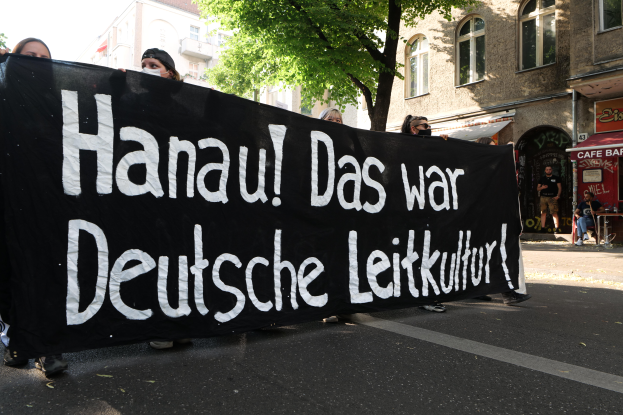 Eine Gruppe von Menschen geht eine Straße entlang und hält eine schwarze Fahne mit der Aufschrift "Hanau das war deutsche Leitkultur" hoch, mit Gebäuden, Bäumen und einem klaren blauen Himmel im Hintergrund.