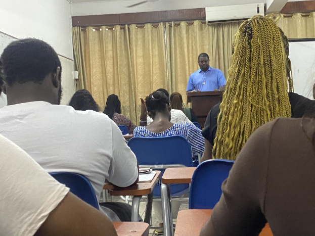 Ein als Gesundheits- und Sozialminister identifizierter Mann steht an einem Rednerpult und spricht zu einer Klasse von sitzenden Menschen, mit Büchern und Gegenständen auf Tischen, einem Vorhang und einer Klimaanlage im Hintergrund.