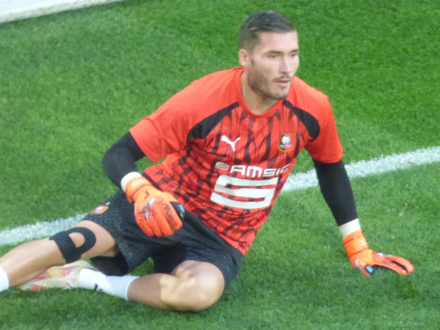 Ein Fußballer in rotem T-Shirt, schwarzen Shorts, weißen Socken und orangen Baseball-Handschuhen sitzt auf dem Boden mit Händen auf den Knien.