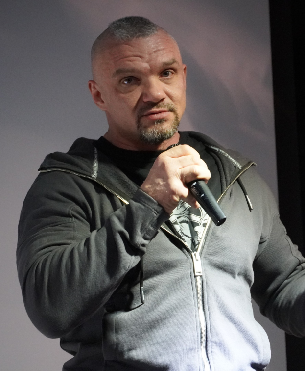 Ein Mann in einer schwarzen Kapuzenjacke und einem schwarzen Anzug mit einem weißen Hemd, der ein Mikrofon hält, steht selbstbewusst vor einer Wand und sieht aus wie ein Komiker mit zurückgekämmten Haaren.