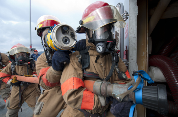 Feuerwehrleute in Schutzausrüstung mit einem Schlauch haltenden, neben einem Mast und Rohren gegen einen bewölkten Himmel stehend.