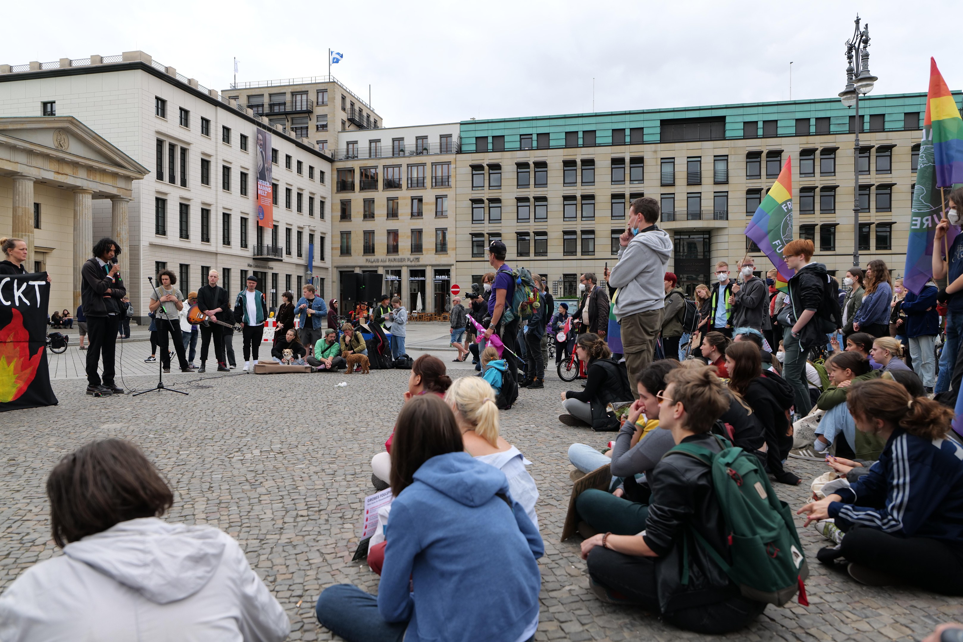 Eine Gruppe von Menschen, die auf dem Boden vor einer Menge sitzen, die Fahnen und Transparente hält, während einer Demonstration gegen Homosexuelle in Berlin, mit einer Statue, Gebäuden und einer Person, die in ein Mikrofon spricht, im Hintergrund.
