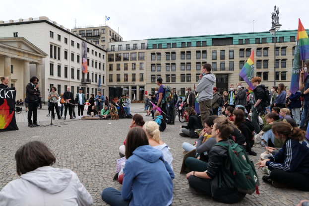 Eine Gruppe von Menschen, die auf dem Boden vor einer Menge sitzen, die Fahnen und Transparente hält, während einer Demonstration gegen Homosexuelle in Berlin, mit einer Statue, Gebäuden und einer Person, die in ein Mikrofon spricht, im Hintergrund.