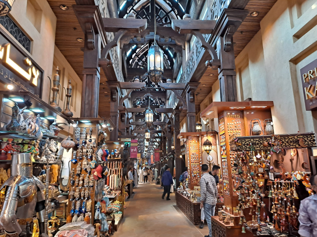 Ein belebter Dubai-Basar mit Menschen, die zwischen Gestellen mit Waren hindurchgehen, beleuchtet von Deckenlampen und Wänden mit Texttafeln.