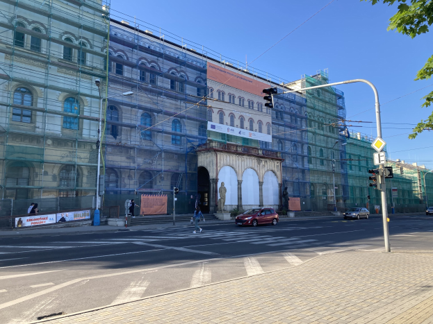 Ein großes Gebäude mit Gerüst, das renoviert wird, umgeben von Fahrzeugen, Passanten, Ampeln, Hinweistafeln, Straßeninfrastruktur, Bäumen und einem klaren blauen Himmel.