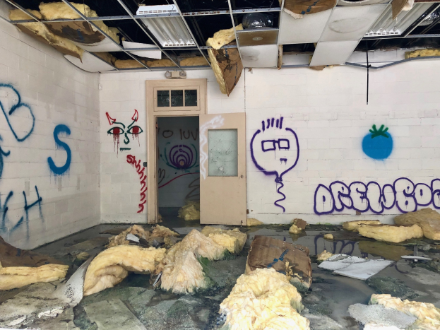 Ein Raum mit Graffiti an den Wänden, eine Tür im Hintergrund, Deckenstangen und Lampen sowie verschiedene Gegenstände auf dem Boden, die tragische Ereignisse im Detroit-Bereich symbolisieren.