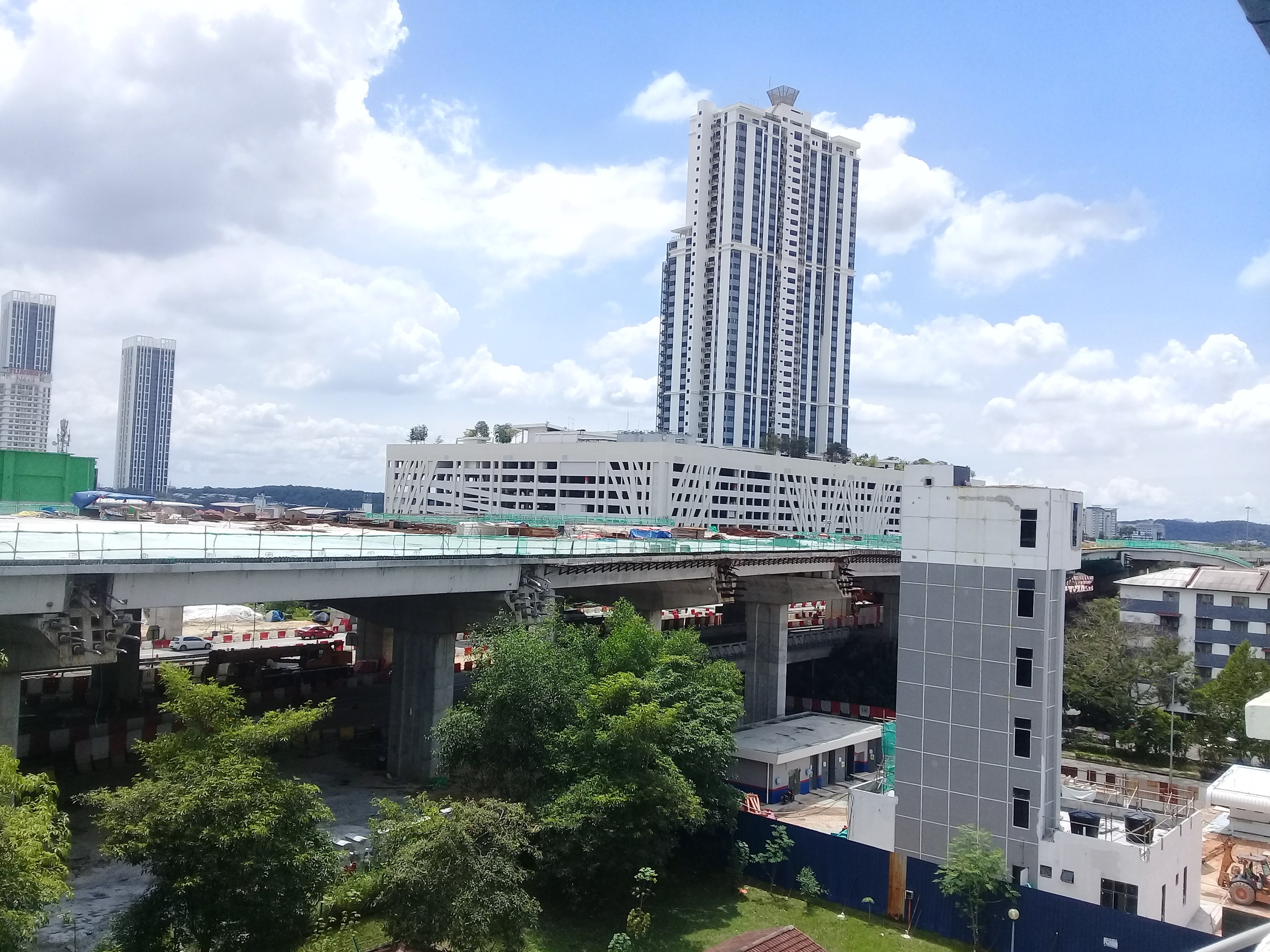 Luftaufnahme einer Stadt mit Gebäuden, Bäumen, Gras, Fahrzeugen, einer Brücke, Masten und einem bewölkten Himmel, die eine Baustelle für einen neuen Wohnkomplex zeigt.