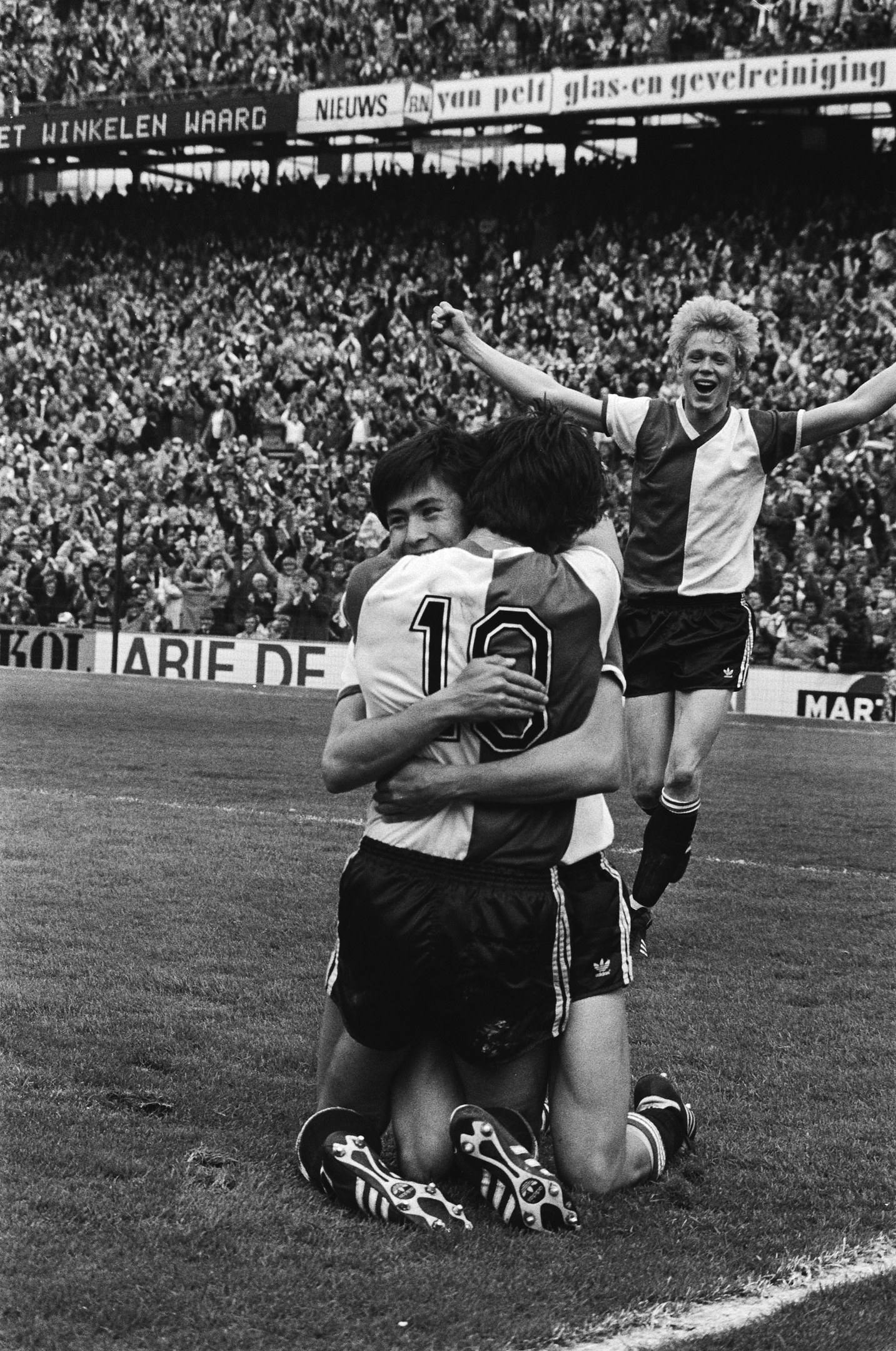 Drei Fußballspieler, die sich nach einem Tor auf dem Boden umarmen, mit jubelnder Menge und schwenkenden Fahnen in einem schwarz-weißen Stadion Setting.