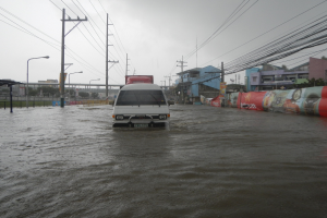 Ein Van fährt durch eine überflutete Stadtstraße mit Strommasten, Laternenmasten und Gebäuden auf beiden Seiten, eine Brücke im Hintergrund und der Himmel oben im Bild.