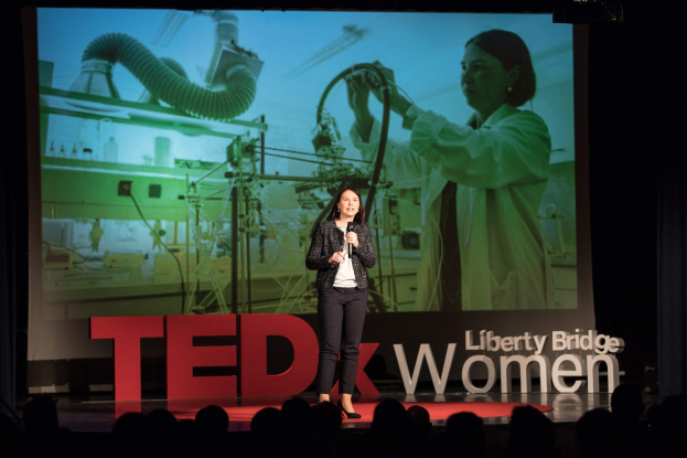 Frau auf einer Bühne mit Mikrofon, im Hintergrund ein Display mit Bild einer Frau und Text zu einem TEDxWomen-Event