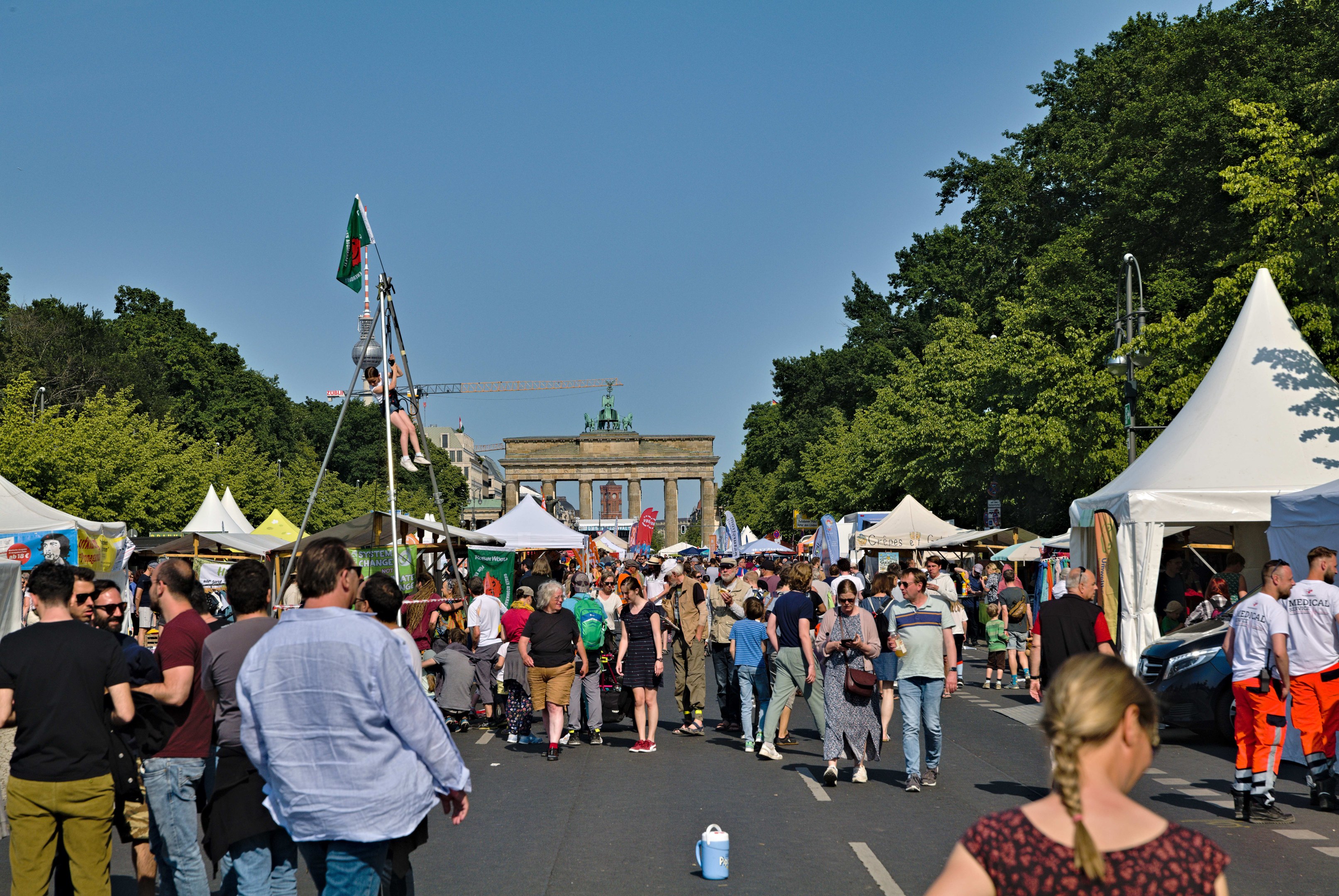 Eine Menschenmenge, die eine Straße mit Zelten, Fahrzeugen und Bäumen entlanggeht, mit einem Bogen und einem klaren blauen Himmel im Hintergrund und Fahnenmästen auf der linken Seite.