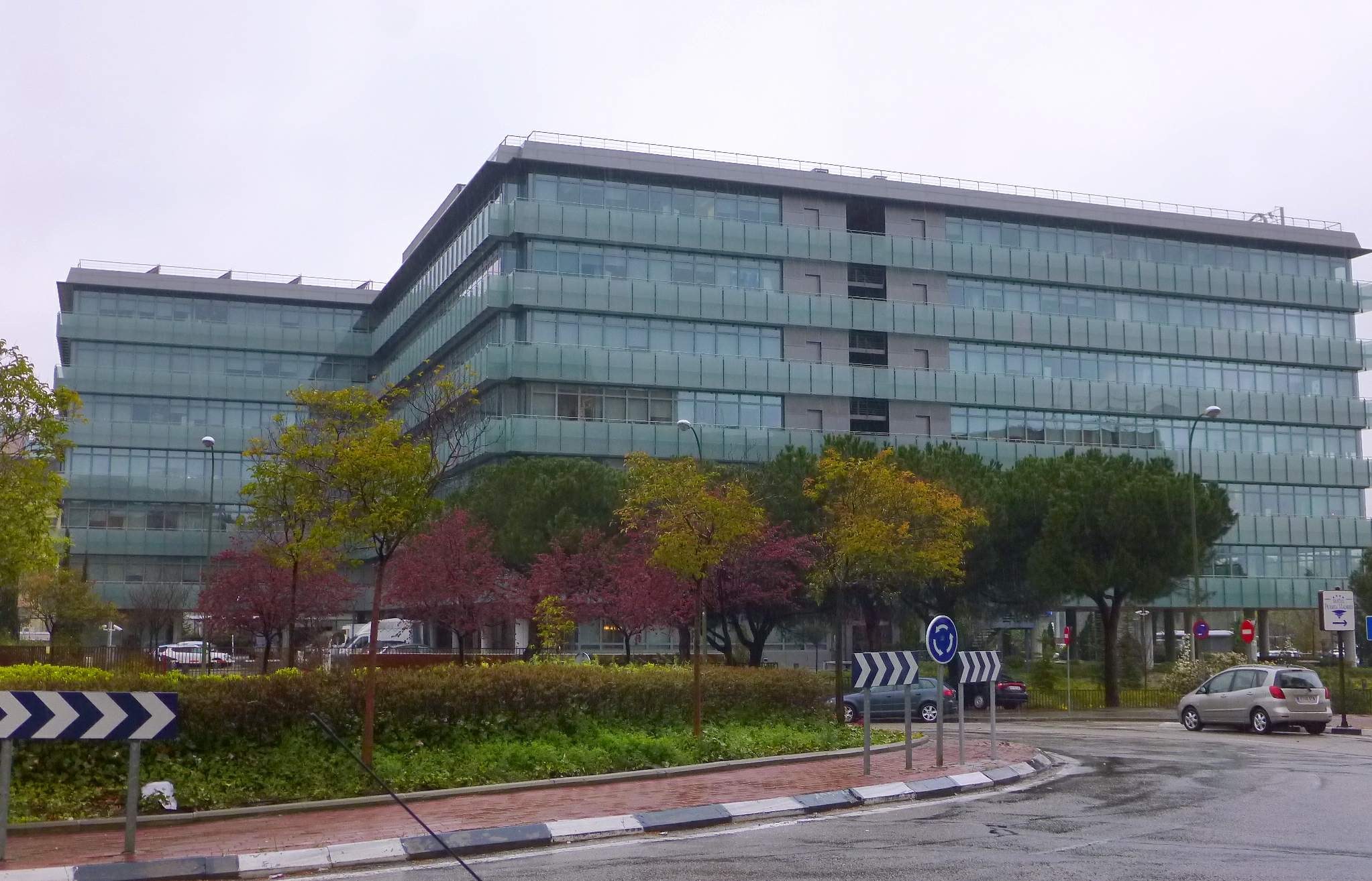 Großes modernes Gebäude mit vielen Fenstern, der Hauptsitz der Europäischen Zentralbank, umgeben von Grünflächen, Straßeninfrastruktur und Fahrzeugen unter einem bewölkten Himmel.