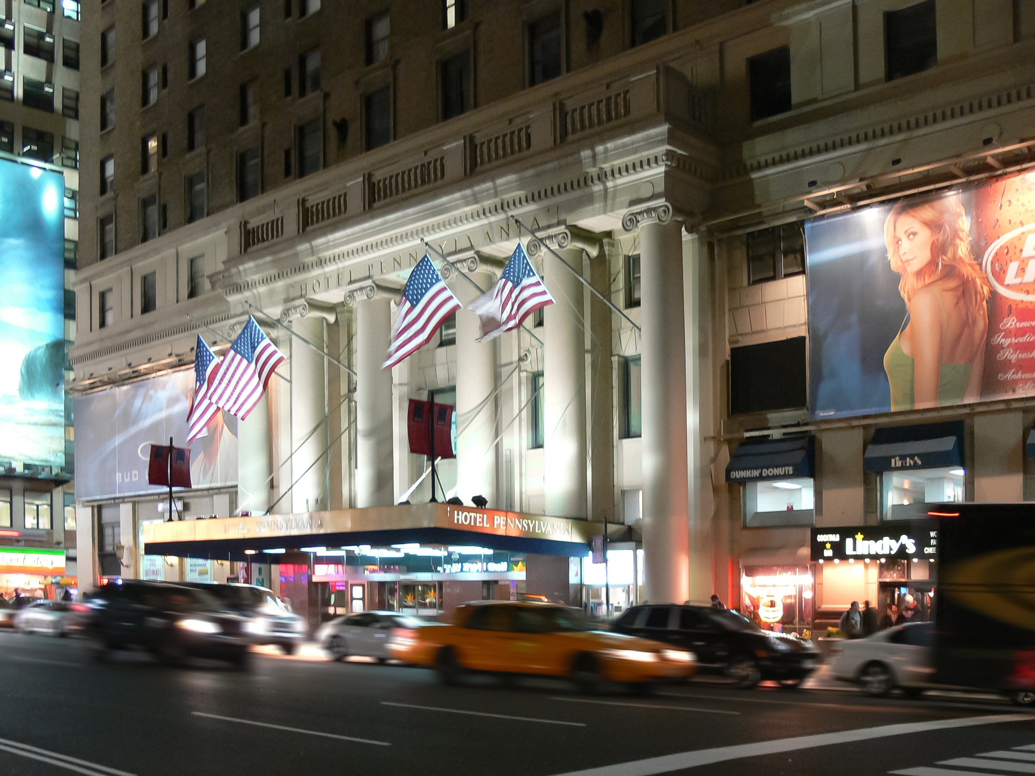 Eine belebte Stra√e in einer Stadt bei Nacht mit fahrenden Fahrzeugen, ein Gebäude mit Fenstern, Pfeilern und Fahnenmasten, beleuchtete Schilder mit Text und ein Bild einer Frau sowie helle Lichter.