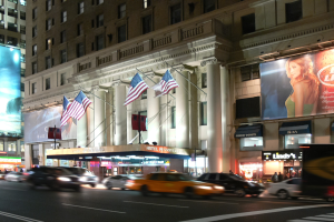 Eine belebte Stra√e in einer Stadt bei Nacht mit fahrenden Fahrzeugen, ein Gebäude mit Fenstern, Pfeilern und Fahnenmasten, beleuchtete Schilder mit Text und ein Bild einer Frau sowie helle Lichter.