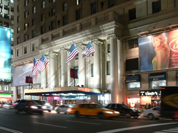 Eine belebte Stra√e in einer Stadt bei Nacht mit fahrenden Fahrzeugen, ein Gebäude mit Fenstern, Pfeilern und Fahnenmasten, beleuchtete Schilder mit Text und ein Bild einer Frau sowie helle Lichter.