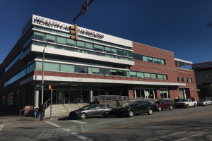 Ein großes Gebäude mit parkenden Autos davor, umgeben von Straßenmasten, Straßenlaternen, Verkehrspfosten, Verkehrsampeln, Schildern, Menschen auf dem Gehweg, Geländern, Treppen, Bäumen und einem klaren blauen Himmel, identifiziert als das neue York City Health Care Obdachlosenzentrum.