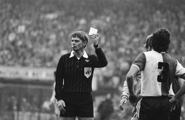 Schwarzes und weißes Bild eines Schiedsrichters, der eine rote Karte hält, mit einer Menge von Menschen in Fußballuniformen dahinter einem Zaun.