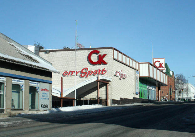 Ein Sportgeschäft in einer Stadt an einer Straßenecke, umgeben von Gebäuden, Straßenlaternen, Bäumen und Schnee, mit dem Himmel im Hintergrund.