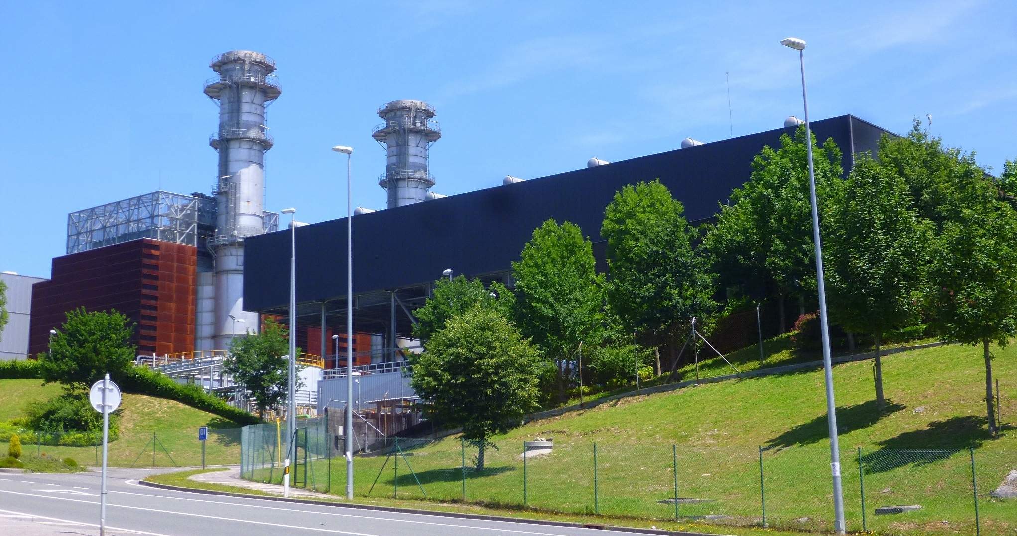 Großes Industriegebäude mit zahlreichen Fenstern, als Kraftwerk identifiziert durch sichtbare Beschriftung, umgeben von städtischer Infrastruktur einschließlich Straßenlaternen, Fahrzeugen und Grünflächen unter einem bewölkten Himmel.