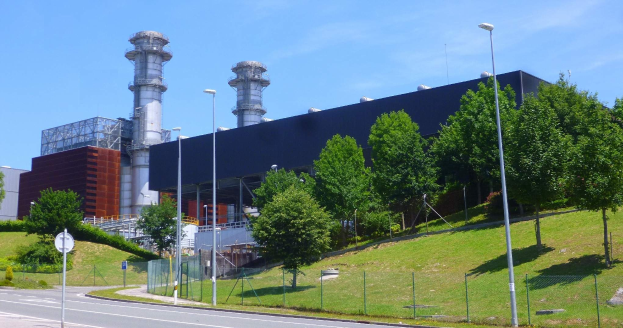 Großes Industriegebäude mit zahlreichen Fenstern, als Kraftwerk identifiziert durch sichtbare Beschriftung, umgeben von städtischer Infrastruktur einschließlich Straßenlaternen, Fahrzeugen und Grünflächen unter einem bewölkten Himmel.